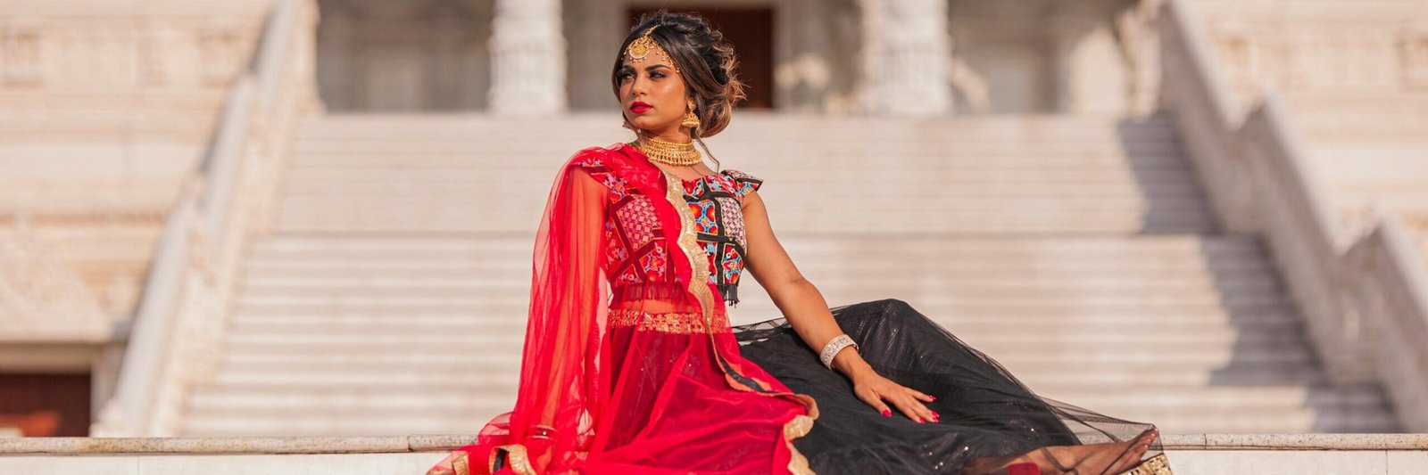 Stylish Indian woman in vibrant red and black traditional dress sitting gracefully on temple steps in Atlanta, GA.